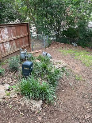 A yard with a wooden fence, utility box, ornamental grasses, and scattered rocks near a wooded area.