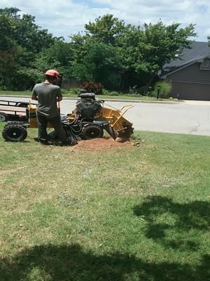 A person in a hard hat operates a yellow stump grinder on a residential lawn, carving into the soil.
