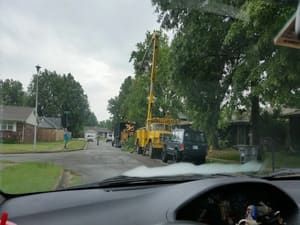A yellow utility truck with an extended boom is parked on a suburban street, with crews working on nearby trees.