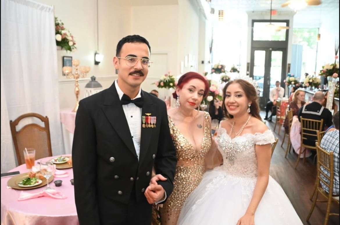 A man in a navy uniform stands with two women in an elegant setting, likely at a wedding reception. The bride wears a white gown, the other woman a gold dress.