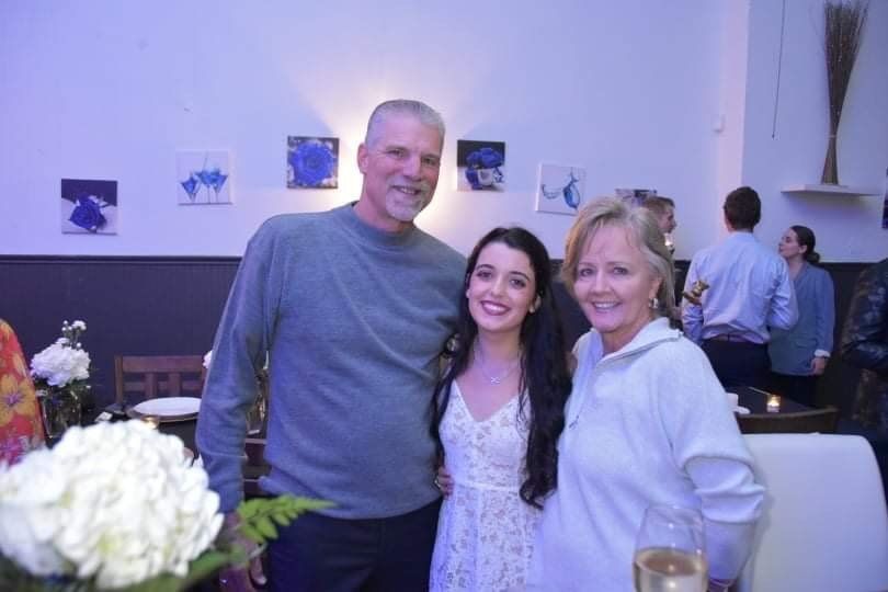 A man and two women smile for a photo inside a restaurant. The woman in the middle wears a white dress, and the others are in casual clothes.