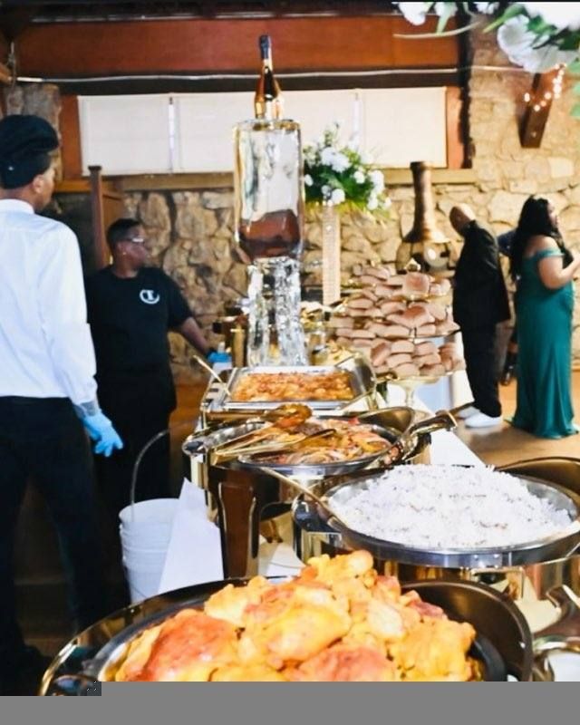 A catering buffet with various dishes and servers. The buffet is set up in front of a stone wall and features an elaborate ice sculpture with a liquor bottle.