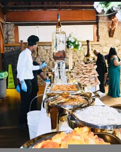 Catered buffet at an event, likely a wedding. A person serves food while others look on. Various dishes are arranged on a long table with an ice sculpture centerpiece.
