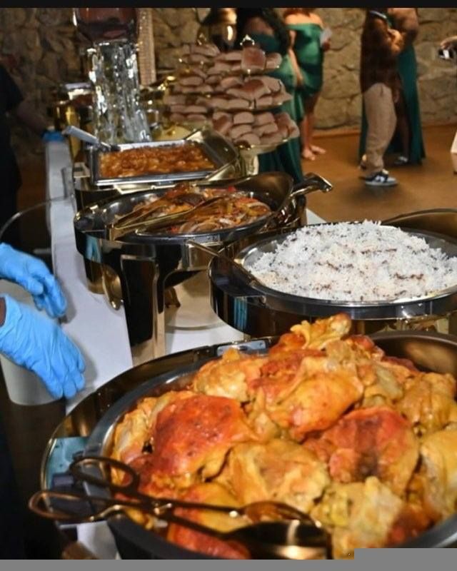 Buffet table at a gathering, with various dishes like rice, stuffed cabbage rolls, and other hot foods in chafing dishes. Someone in blue gloves is present.