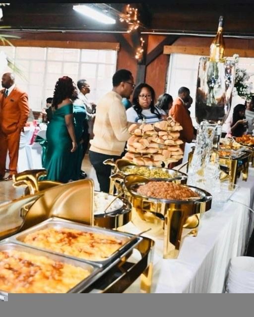 A buffet-style spread with various dishes and people gathering around at an event.  Guests in formal attire are selecting food.