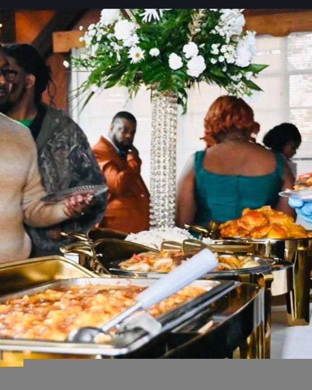 People at a buffet, selecting food. A floral centerpiece stands behind the food.