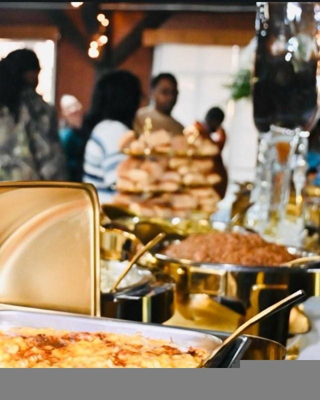 Buffet table with gold serving dishes and a crowd of people in the background. Various foods are visible.