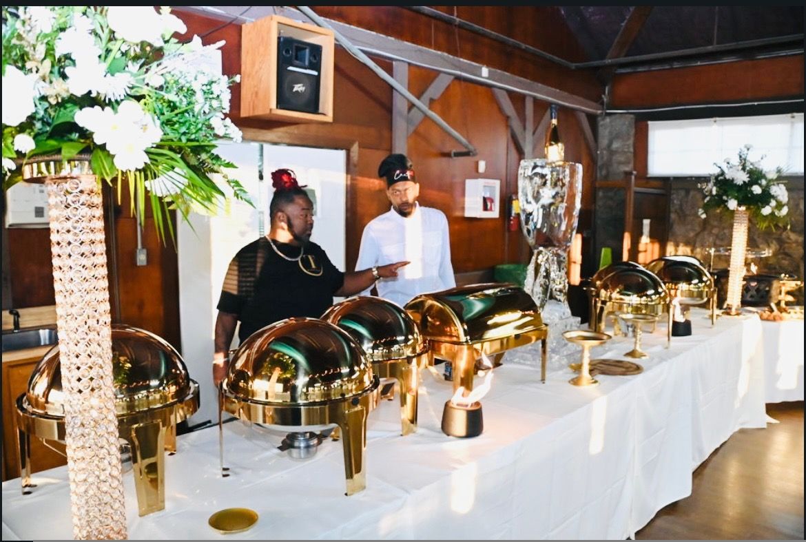 A buffet table with golden serving dishes and floral arrangements. Two people, a woman and a man, stand behind the table.