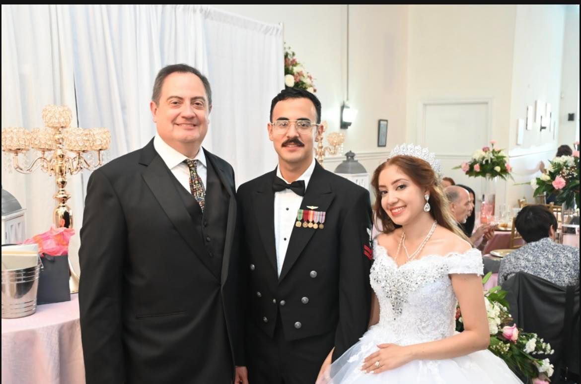 Three people pose for a photo at a wedding. A man in a tuxedo stands left, the groom in his Navy uniform in the middle, and the bride in a white gown on the right, all smiling.