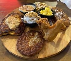 A round wooden board with various breakfast foods: french toast, pancakes, waffles, eggs, fried chicken, and a side of grits.