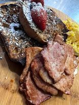 French toast with powdered sugar and strawberries, next to bacon and scrambled eggs on a wooden board.