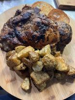 Grilled pork chop with roasted potatoes and rolls on a wooden serving board. The pork chop is brown and charred.