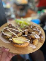 A wooden plate holds a burger, meat, and lettuce, drizzled with sauce. A hand holds the plate indoors.