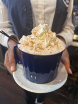 Person holding a blue mug of frothy drink topped with whipped cream and cinnamon, on a saucer.