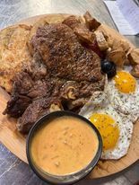A hearty meal on a wooden platter: steak, fried eggs, potatoes, bread, and a bowl of creamy sauce.