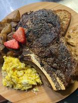 A hearty breakfast plate with a large steak, scrambled eggs, potatoes, toast, and strawberry slices on a wooden board.