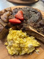 Close-up of a steak with potatoes, scrambled eggs, and strawberries on a wooden plate. The steak is cooked with a dark crust.