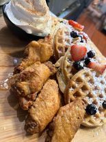 Chicken and waffles on a wooden board. Fried chicken wings beside waffles topped with berries and powdered sugar, and a dish of whipped cream.