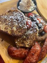 French toast with berries and sausage on a wooden board, garnished with whipped cream and powdered sugar.
