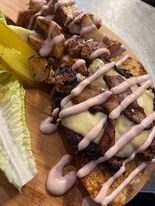 A grilled sandwich on a wooden board with potatoes, pickles, and lettuce. Drizzled sauce and cheese cover the sandwich.