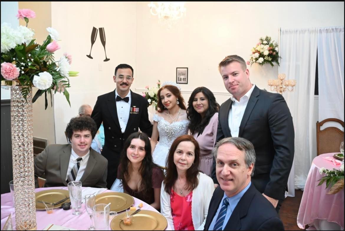 A group of people at a wedding reception, smiling at the camera. The bride and groom are in the center. Tables and floral arrangements are visible.