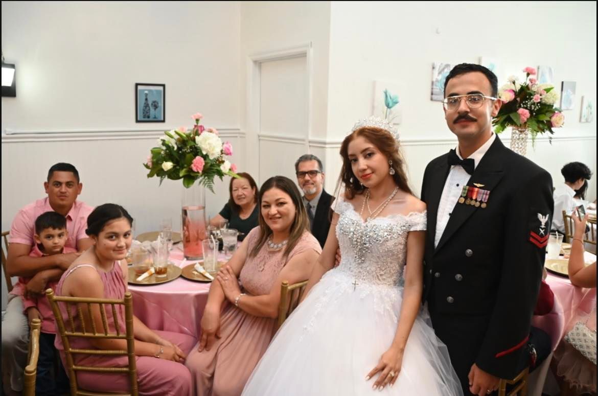 Family poses for a photo at a celebration. A young woman in a white gown stands next to a man in a military uniform. Others are seated at a table decorated with pink.