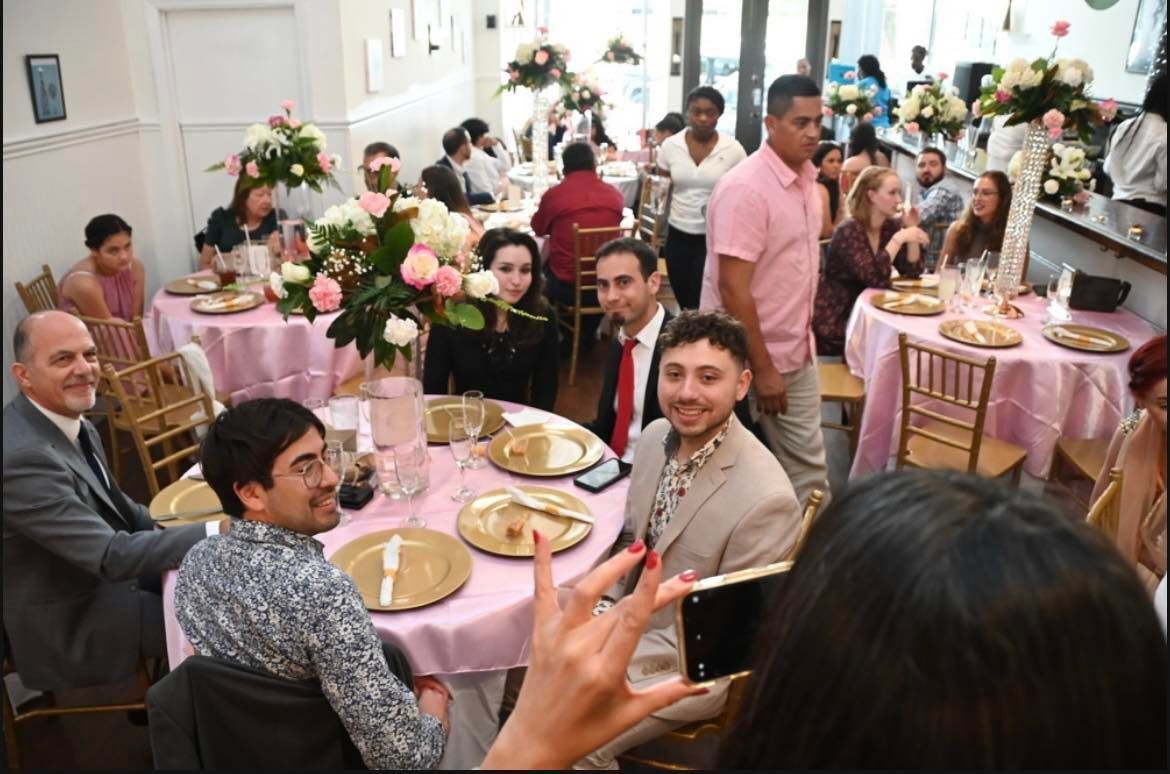 People seated at round tables decorated with pink tablecloths and flowers, celebrating an event indoors. Some are smiling, and a server is present.