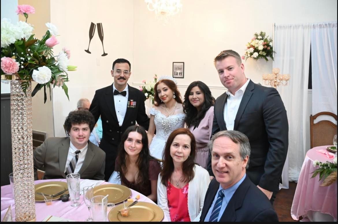 Group photo at a decorated event. People are smiling; a central woman in a white dress appears to be a bride.