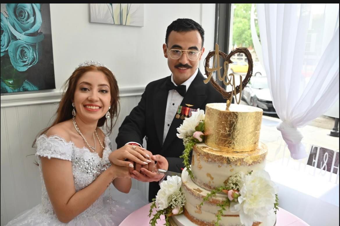A smiling bride and groom cut a three-tiered wedding cake adorned with gold leaf, flowers, and a heart topper. They are indoors, likely at their reception.