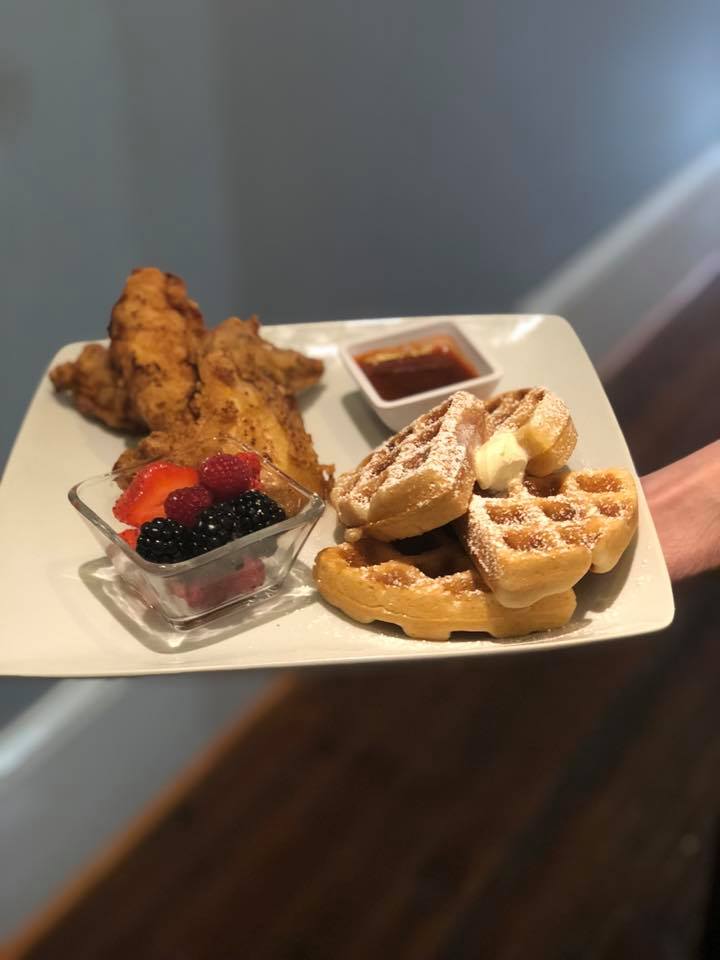 Plate with fried chicken tenders, waffles with butter and powdered sugar, fruit, and syrup.