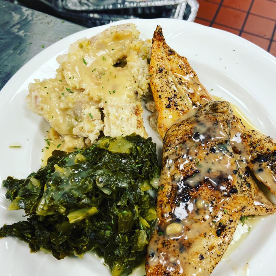 Plate of Southern-style food: grilled fish fillet, creamy grits, and collard greens, all drizzled with gravy.