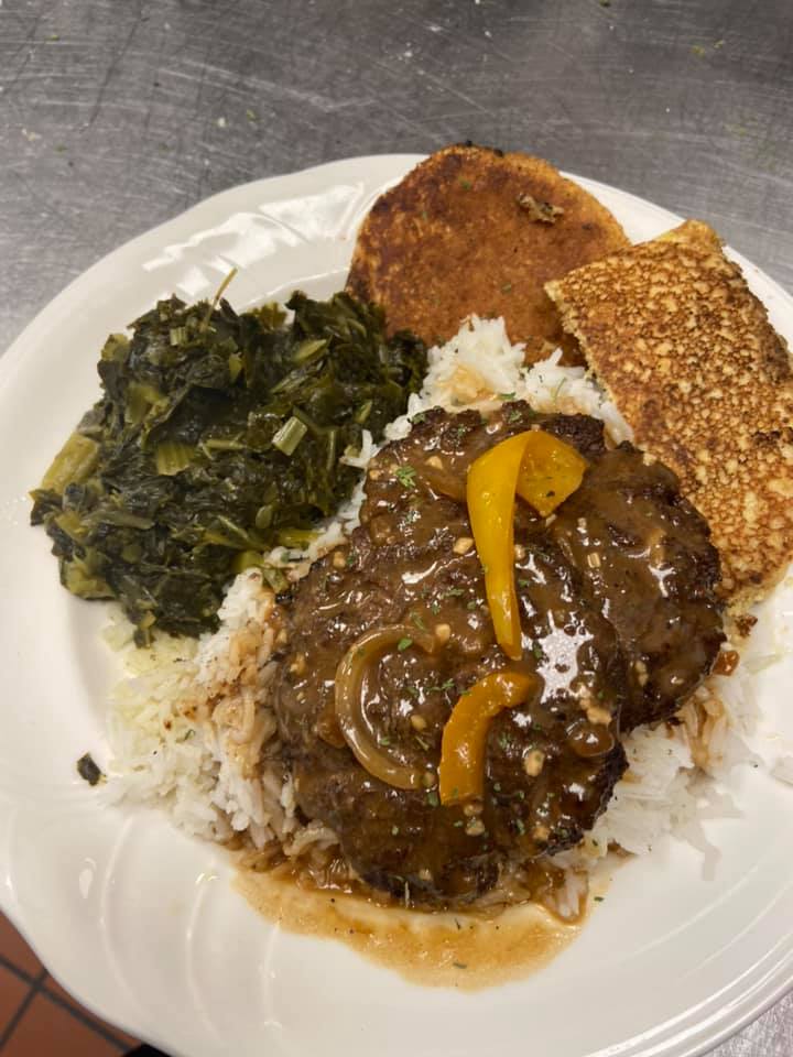 Plate of Southern comfort food: Salisbury steak with gravy, collard greens, rice, a fried patty, and a crispy cornbread square.