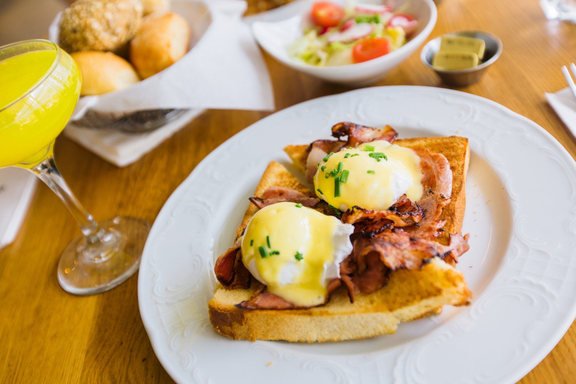 Eggs Benedict on toast with bacon, a side salad, bread rolls, and a glass of orange juice on a wooden table. Eggs Benedict on toast with bacon, a side salad, bread rolls, and a glass of orange juice on a wooden table.