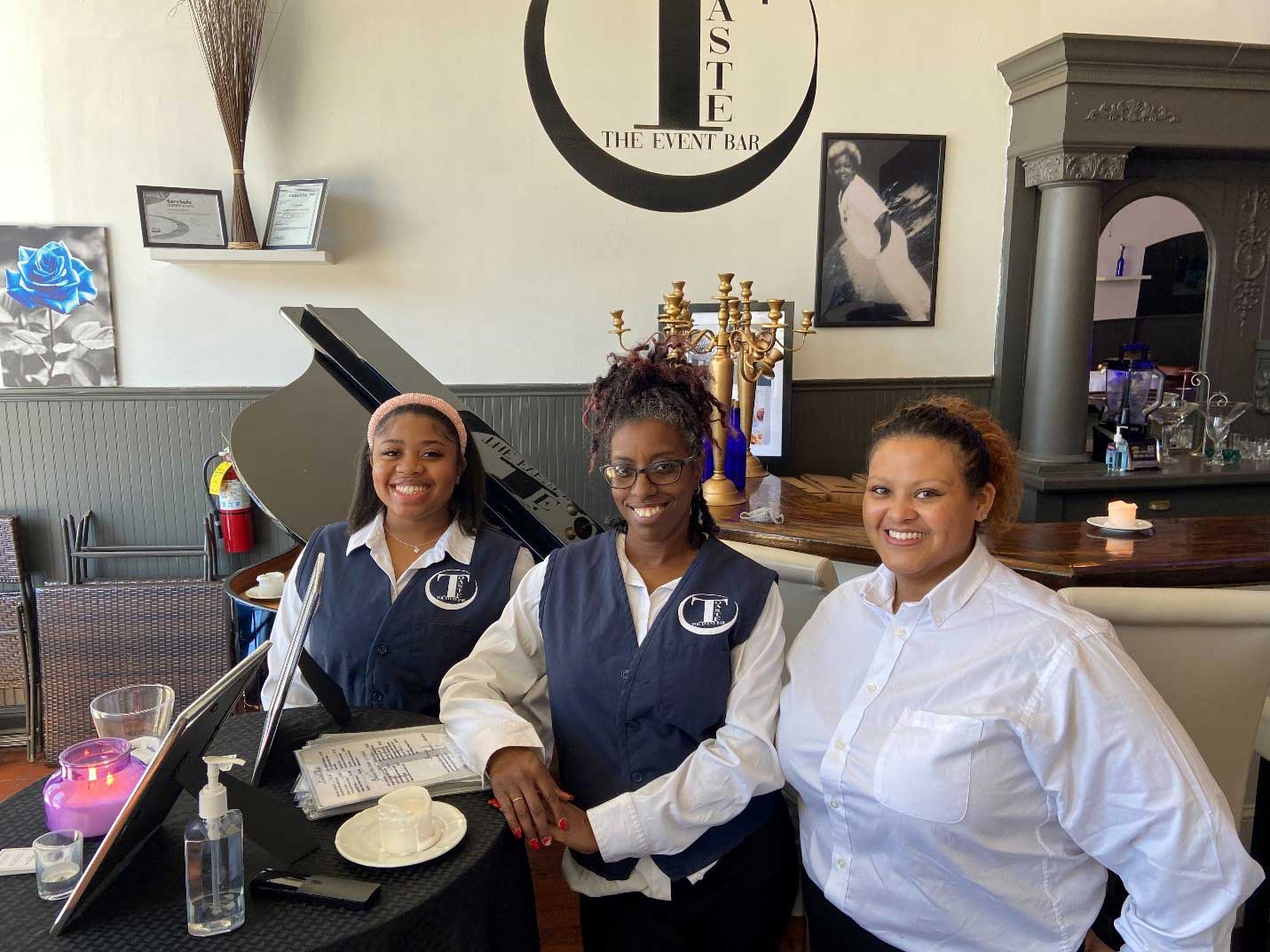 Three women staff members smiling behind a desk. They are wearing matching navy vests over white shirts, and the restaurant has a grand piano and decorative elements.