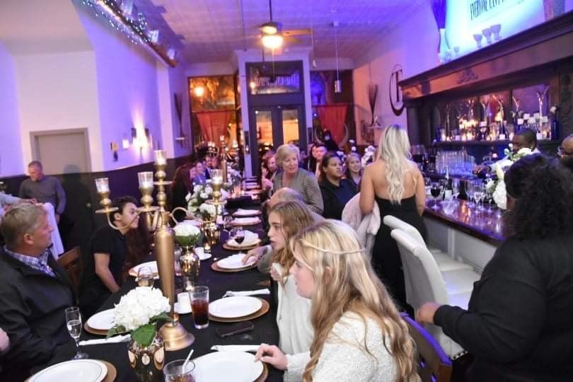 Inside a restaurant, people are seated at a long table set for a formal dinner. Some stand near the bar, others are walking or talking.
