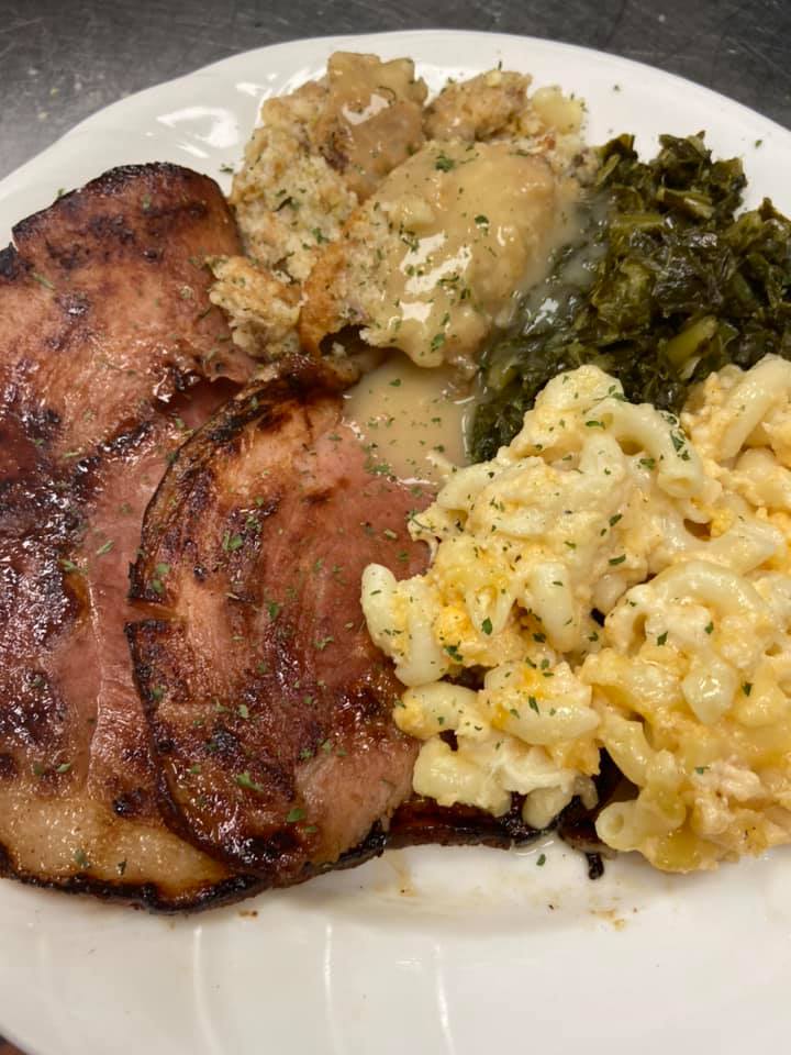 A plate of Southern comfort food featuring sliced ham, mac and cheese, collard greens, stuffing, and gravy.