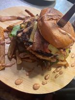 A large burger with fries on a wooden plate, held together by a knife. Pink sauce is drizzled on the plate.