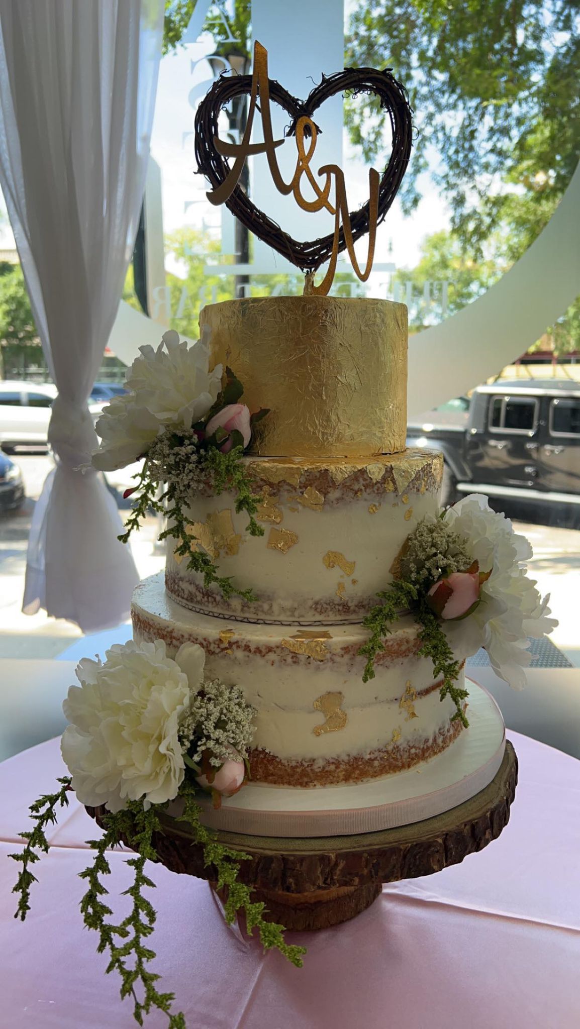 Three-tiered wedding cake with gold accents and floral decorations, topped with a heart-shaped initial topper on a wooden cake stand.
