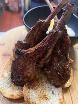 Grilled lamb chops on toasted bread, served with a small bowl of white sauce on a wooden board.