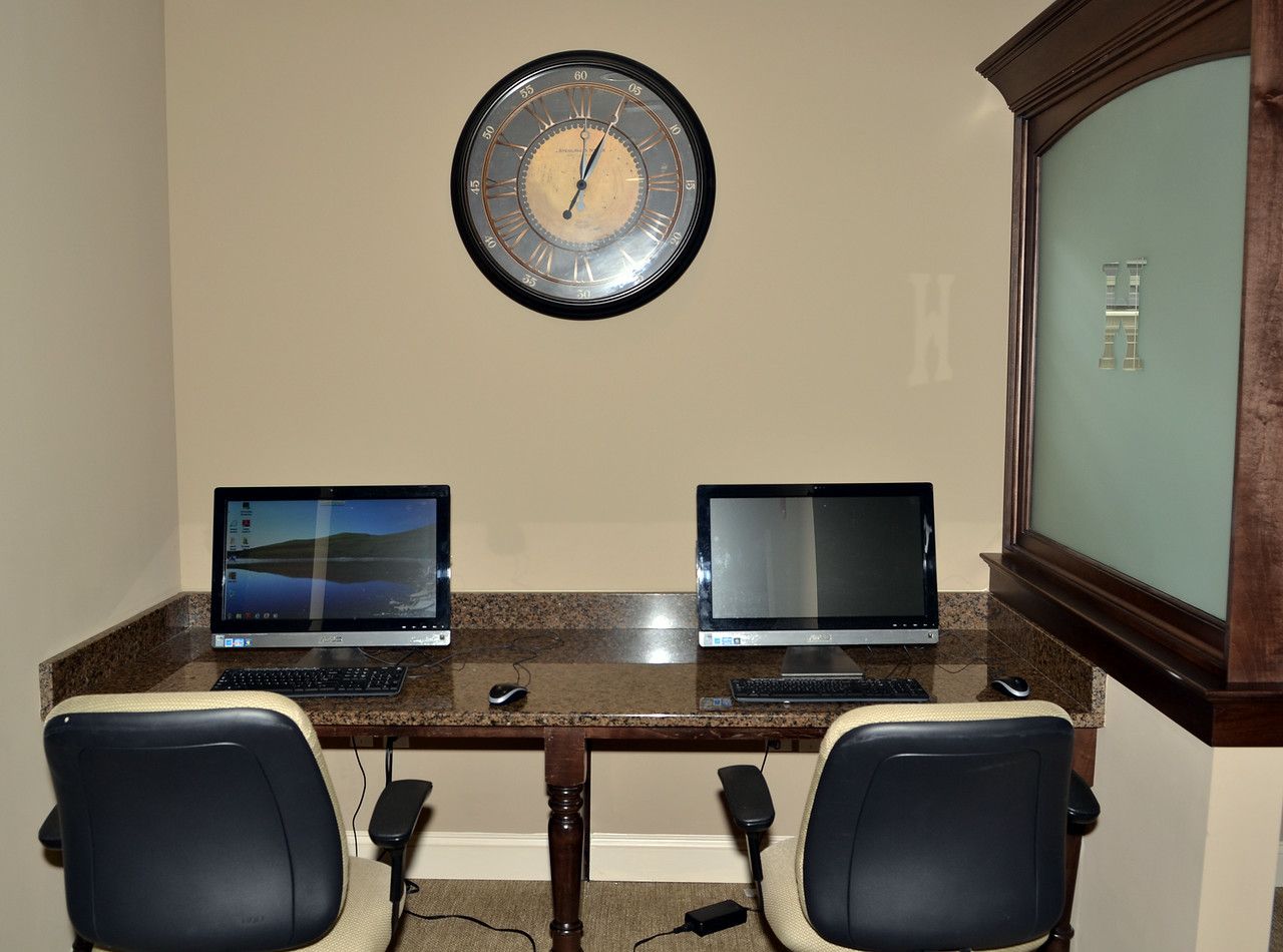 Two computer workstations on a granite counter in a small office area, clock on the wall.