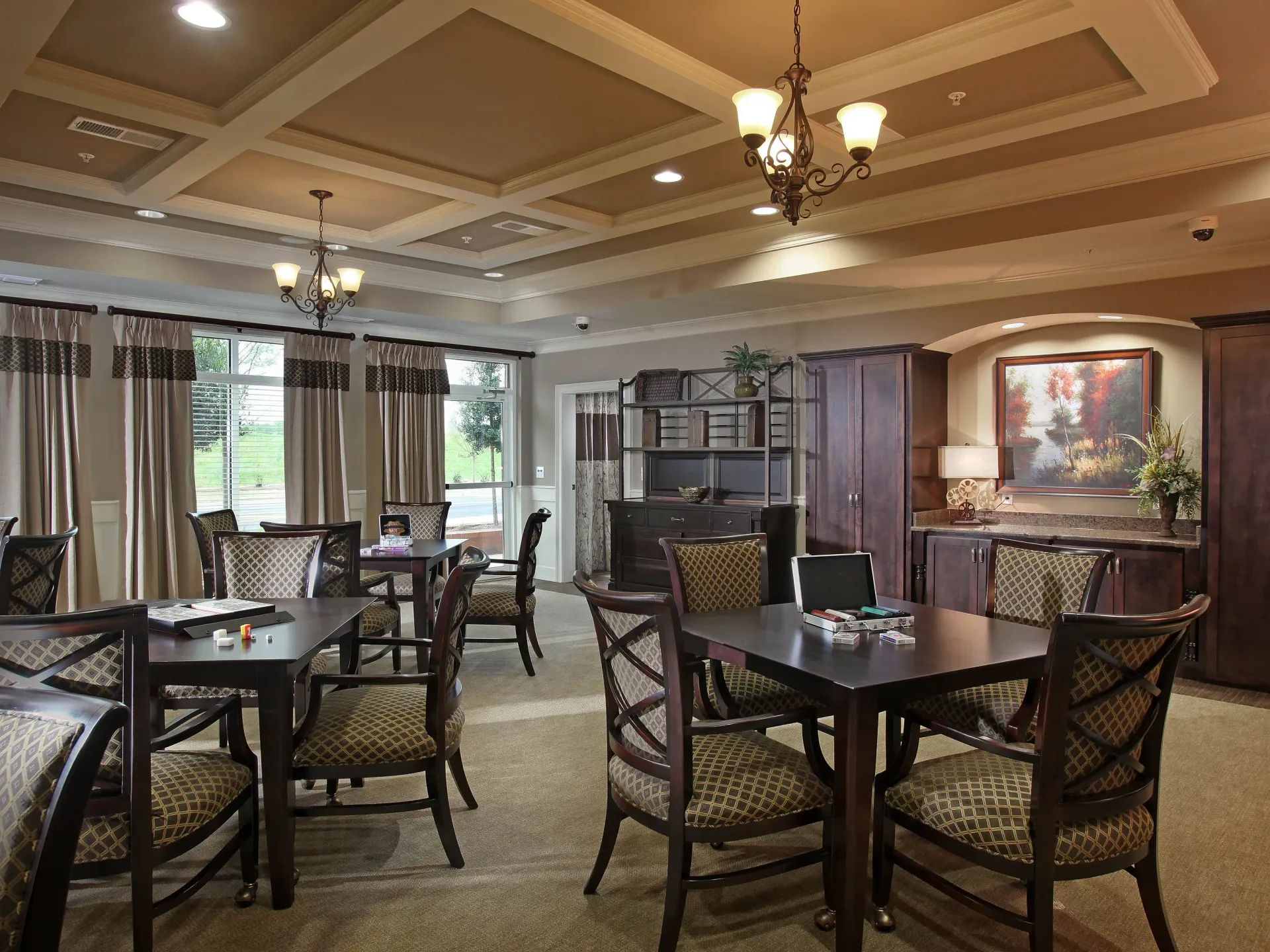 Communal lounge with dark wood tables, upholstered chairs, large windows, and built-in cabinets.