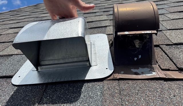 Hand holding a new metallic vent cover next to an old brown vent on a shingled roof.