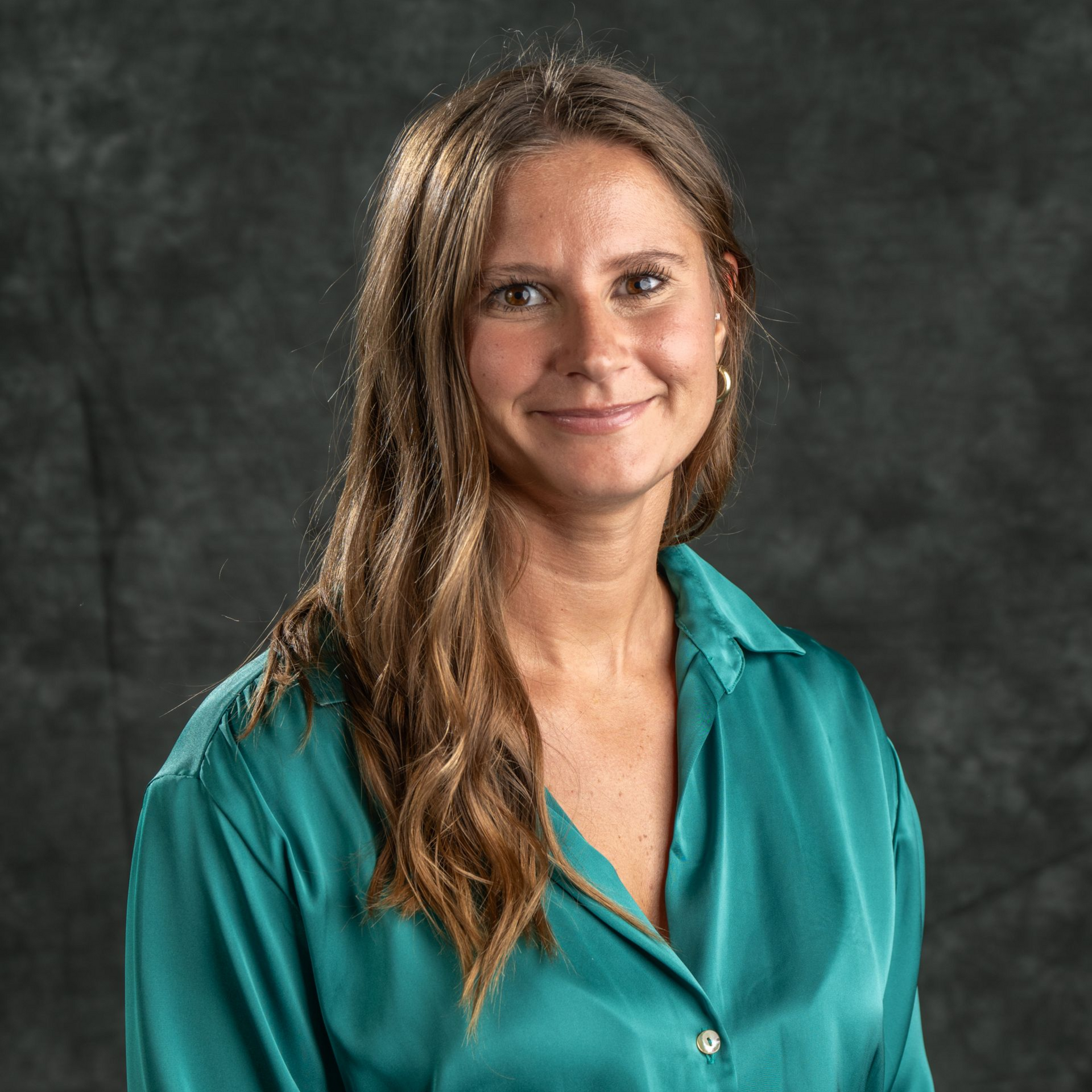 Woman with long brown hair wearing a teal blouse smiling, posing against a dark gray backdrop.