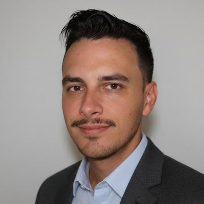 Man in a blue shirt and grey suit jacket, looking at the viewer. He has dark hair, a mustache, and a neutral expression.