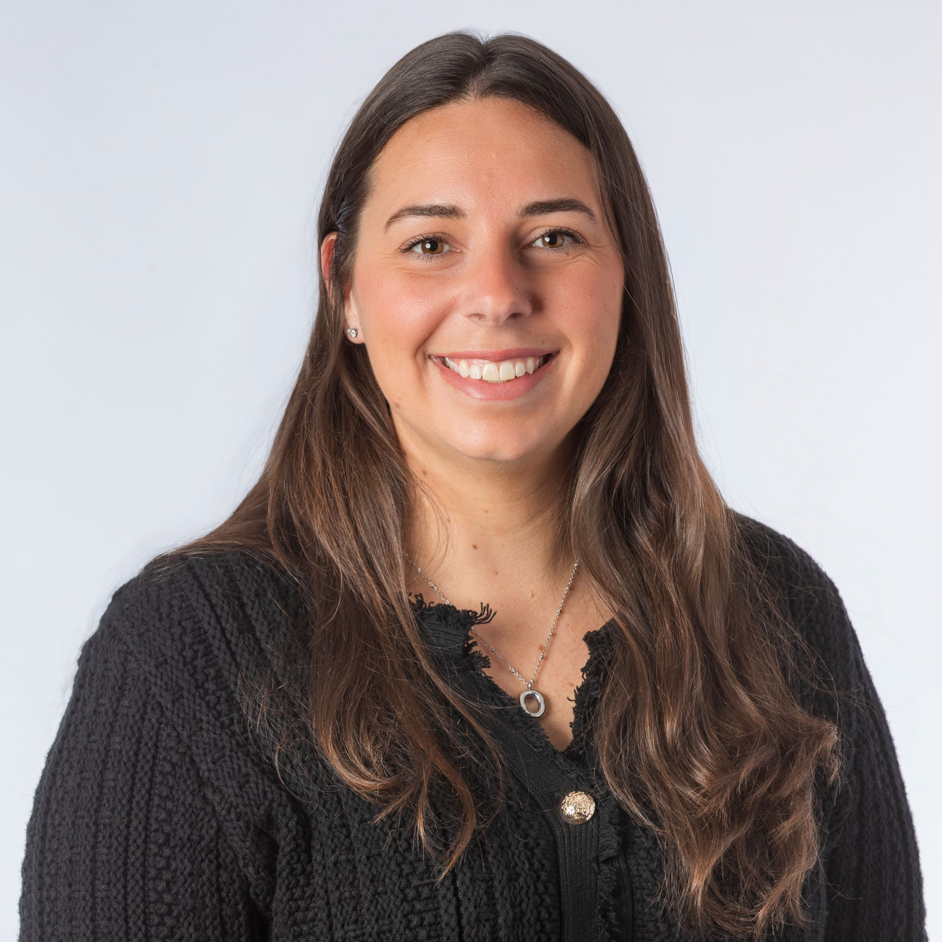 Woman with long brown hair, smiling, wearing a black sweater with gold buttons against a white backdrop.