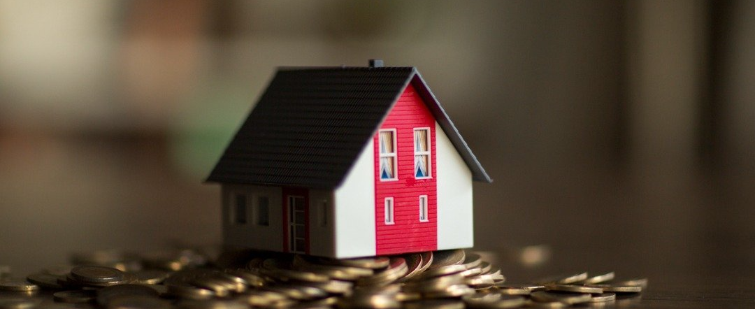 Miniature house with red accents, atop a pile of coins. Blurred background.