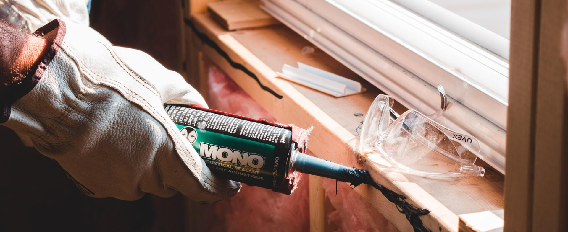A person wearing gloves, applying caulk to a window frame with a caulk gun.