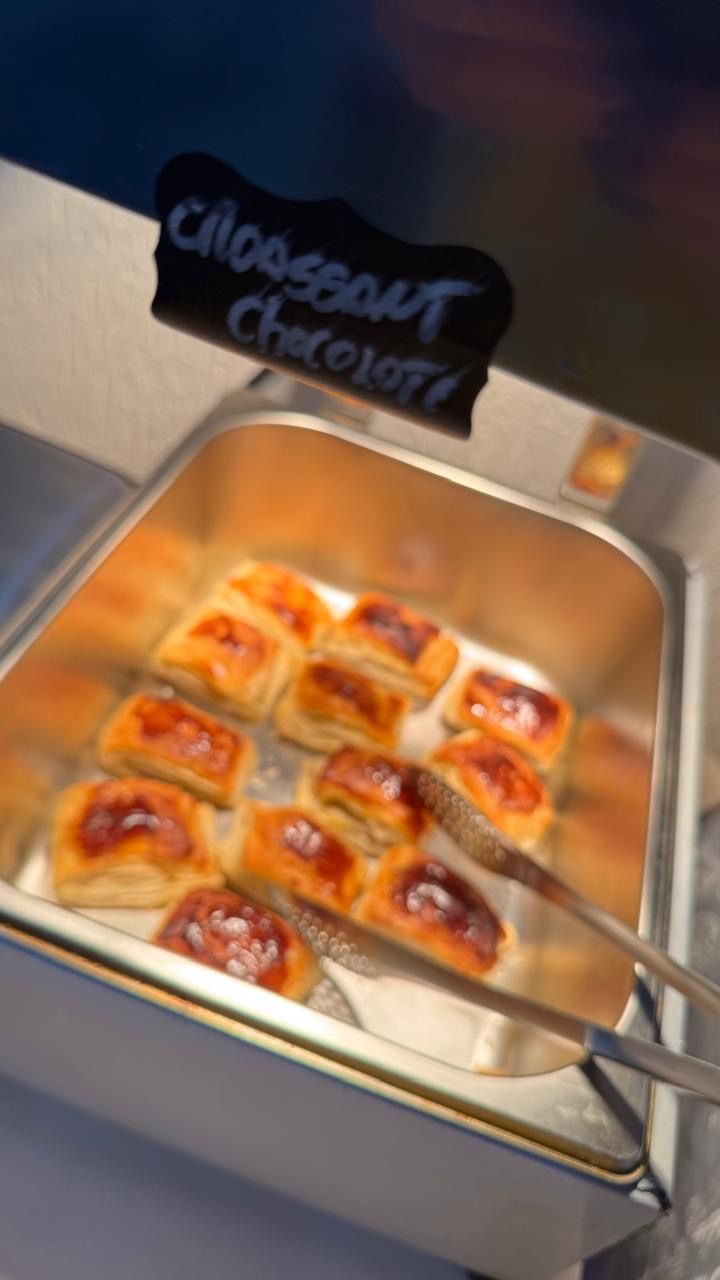 Bandeja de croissants dourados em uma panela de metal com pinças, etiquetada como “croissant de chocolate”.
