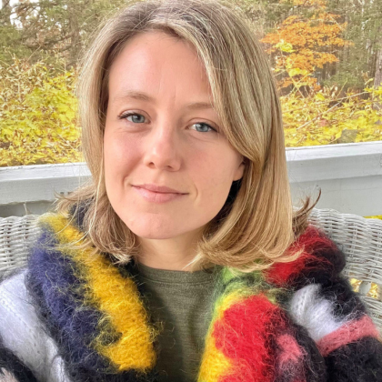 Person with blonde hair wearing a colorful fuzzy sweater on a balcony with autumn trees in the background