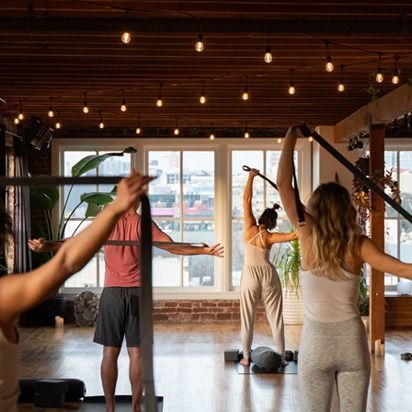 People stretching in a sunlit yoga studio with wooden floors and hanging lights